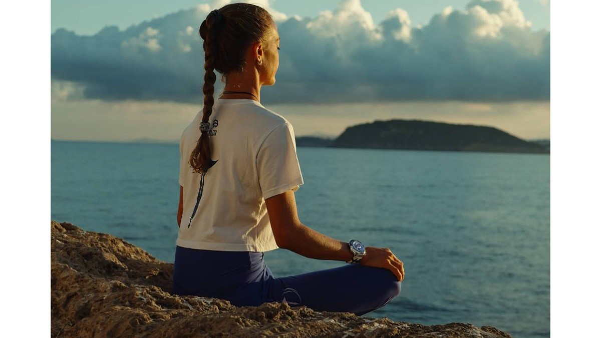 Huawei - poza cu o femeie meditanad la malul oceanului. Are parul saten, prins intr-o coada impletita, sta cu picioarele incrucisate si poarta un tricoul alb si colanti bleumarin