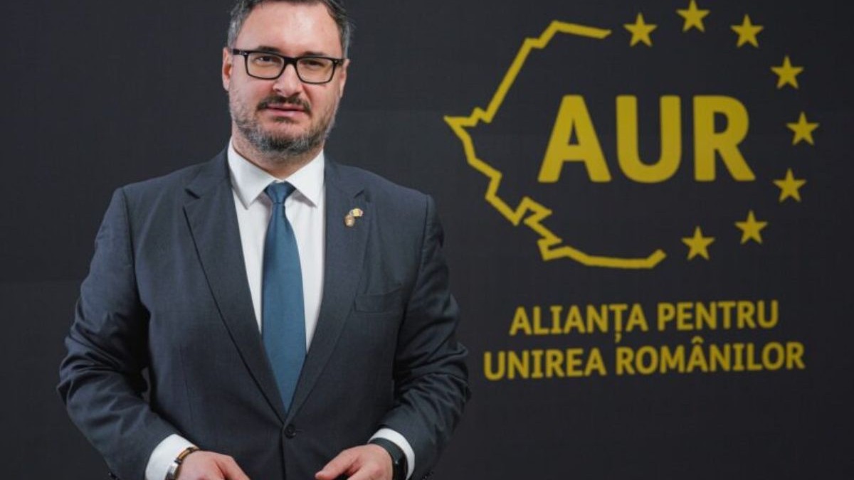 Dan Tanasă, îmbrăcat într-un sacou negru, cămașă albă și o cravată bleumarin, stând cu mâinile pe lângă corp în fața unui fundal negru cu stema aurie a partidului AUR
