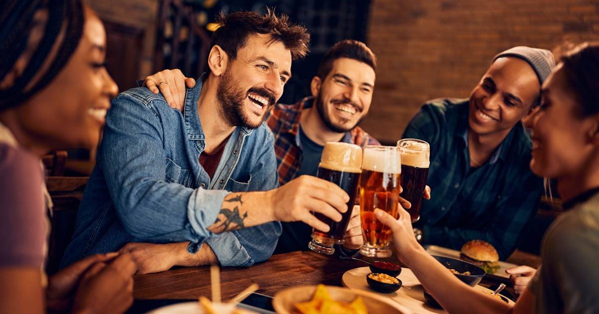 Mai multi tineri la o masa, intr-un pub, rad, se distreaza, dau noroc cu pahare pline cu bere
