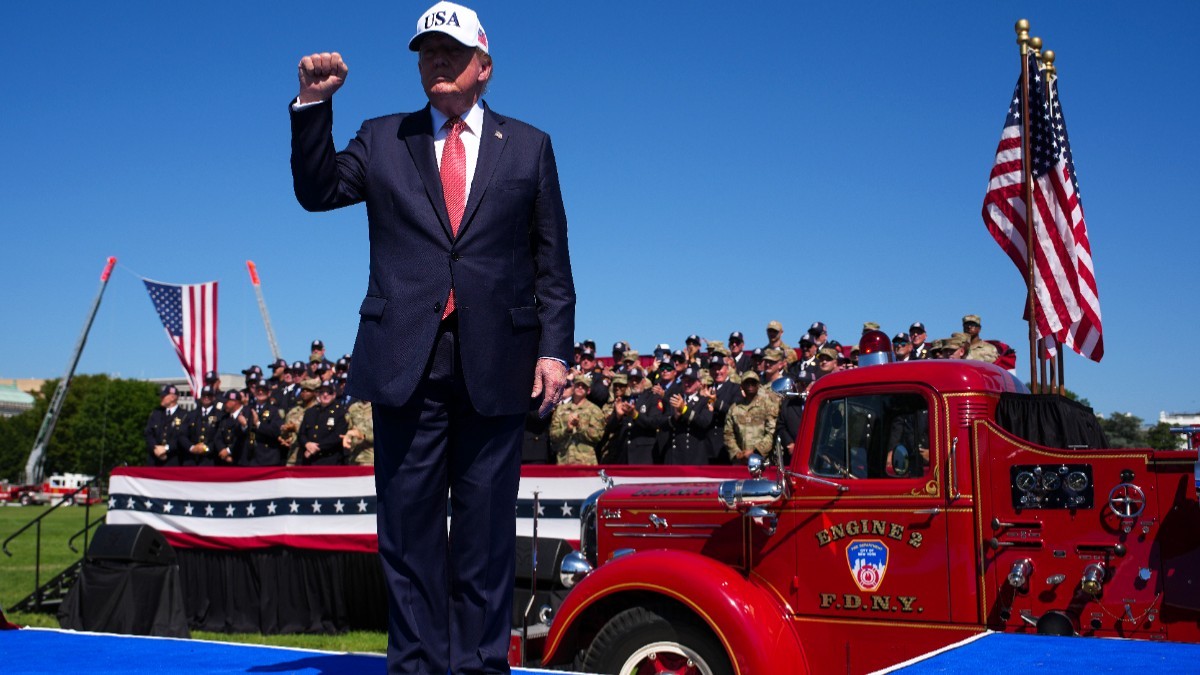 Donald Trump purtând o șapcă albă cu inscripția „USA” și ridicând pumnul, stând în fața unei mașini de pompieri roșii și a unui grup de oficiali în uniformă, pe fundalul unui cer senin.