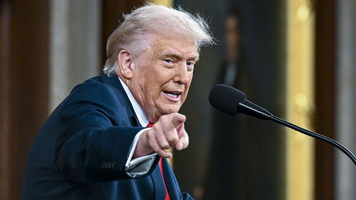 Președintele Donald Trump, îmbrăcat într-un costum bleumarin și cravată roșie, arătând cu degetul spre aparatul de fotografiat, în fața unui microfon.