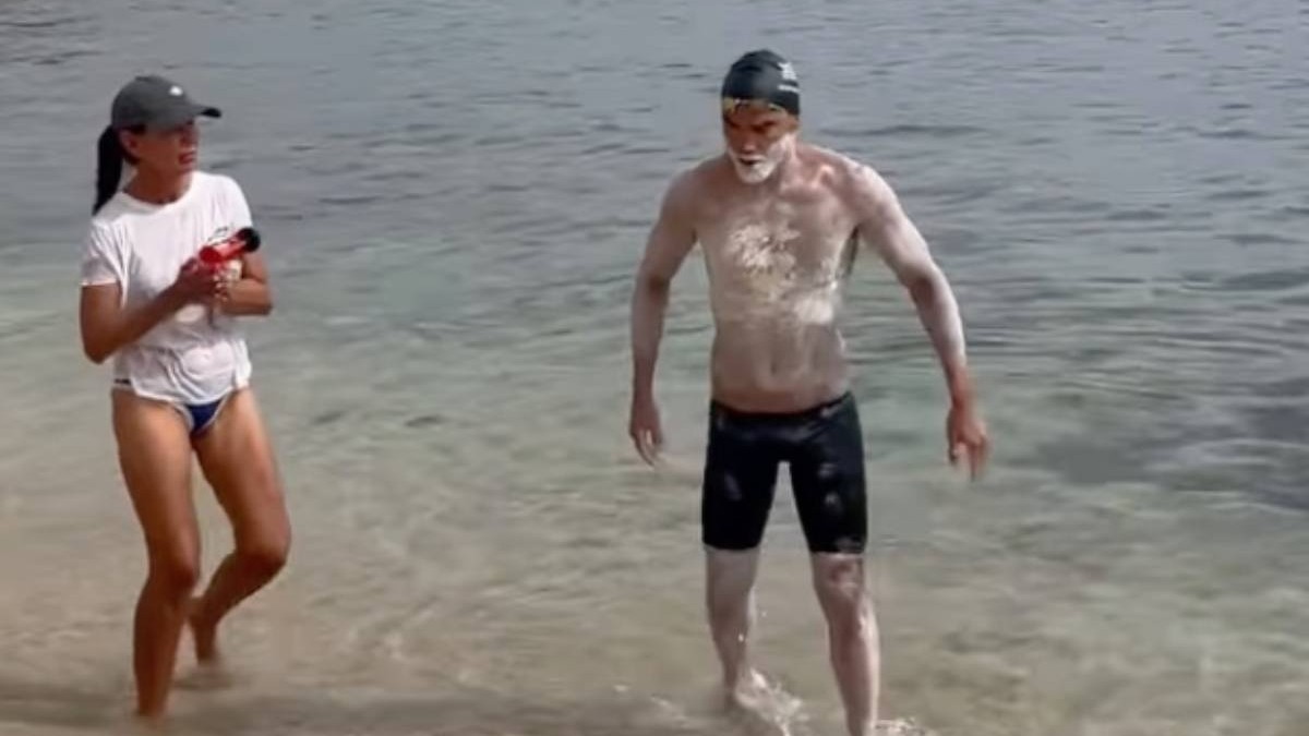 Spyros Chrysikopoulos iese din mare la capătul traversării dintre Grecia și Italia, după aproape 32 de ore de înot. Sportivul poartă cască și ochelari de înot, iar corpul îi este acoperit de cremă de protecție. Lângă el se află o femeie, pe mal, în timp ce acesta pășește prin apa mică spre plajă.