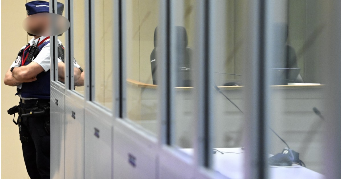 Un ofițer de poliție cu brațele încrucișate și fața blurată stând de pază în stânga unei cabine securizate cu geamuri transparente, în interiorul unei săli de judecată.