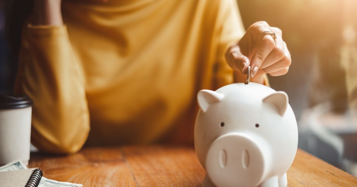 O femeie pune bani într-o pușculiță de tip porc alb, aflată pe o masă. Foto cu caracter ilustrativ: Shutterstock