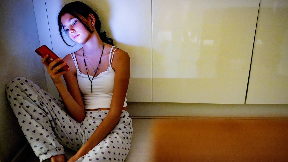 O adolescentă plictisită privește ecranul unui telefon, ea stă pe jos, într-o bucătărie, sprijinită de un dulap