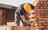 Un muncitor în construcții care poartă cască galbenă de protecție și mănuși, lucrând la zidirea unui perete din cărămidă roșie pe un șantier.