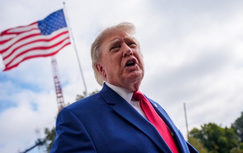 Donald Trump vorbind dintr-un unghi coborât, îmbrăcat în costum albastru și cravată roșie, pe fundalul unui steag al Statelor Unite care flutură în aer liber.