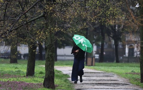 O femeie care are o umbrelă verde pe scrie OK merge pe o alee din parc, într-o zi cu ploaie, iar în jurul ei sunt copaci și iarbă pe jos, București, 3 aprilie 2026