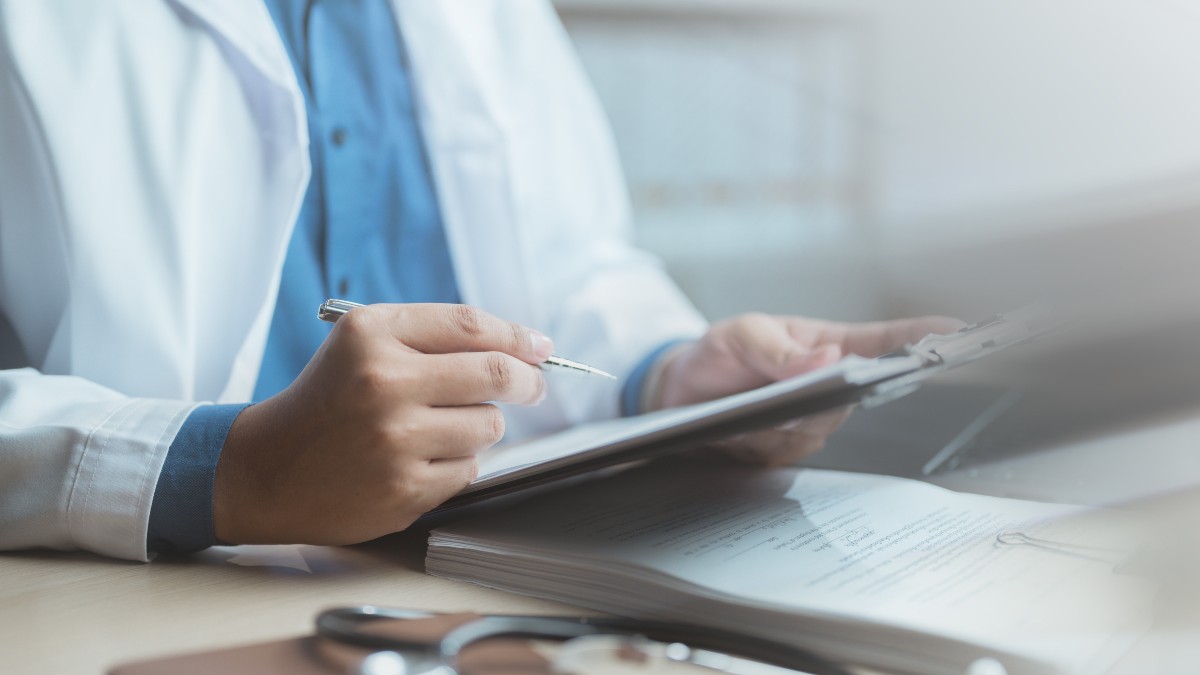 Un doctor care poartă halat alb și stă la birou în timp ce ține un pix și foi în mâini