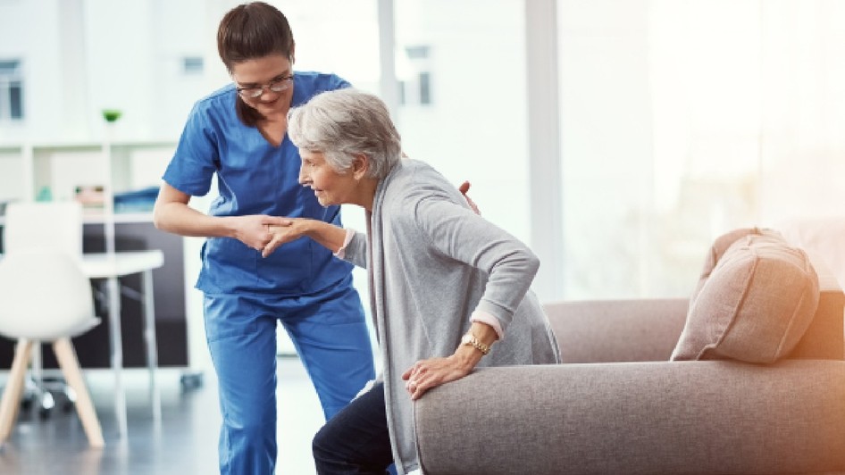 O asistentă medicală o ajută pe o femeie în vârstă să se ridice de pe o canapea gri