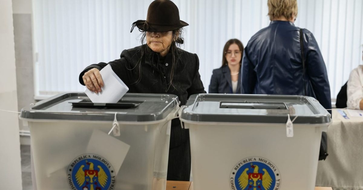 În prim-plan, o femeie în vârstă îmbrăcată în negru și cu o pălărie neagră care îi acoperă fața introduce buletinul de vot în urnă, după ce și-a exprimat opțiunea la alegerile parlamentare din Republica Moldova, iar în spate se mai văd câteva persoane, 28 septembrie 2025