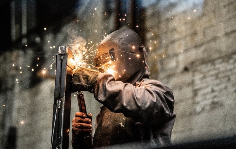 Un sudor purtând mască și echipament de protecție în timp ce sudează o structură metalică, împrăștiind scântei luminoase într-un mediu industrial.