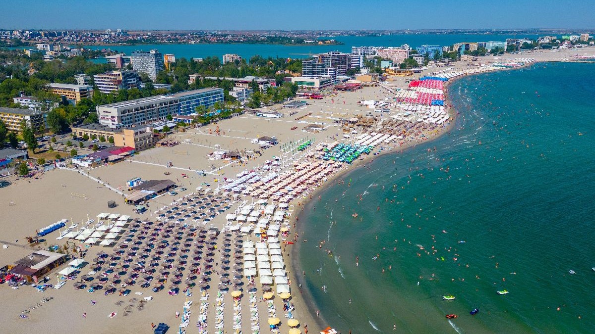 Imaginea prezintă o vedere aeriană a unei plaje aglomerate de pe litoralul românesc, cu numeroase umbrele colorate, hoteluri în fundal și turiști sub un cer senin.