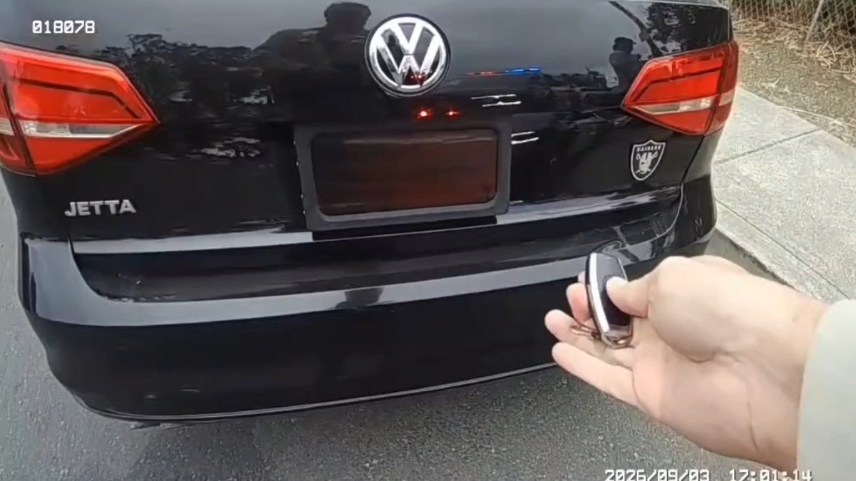 Partea din spate a unui autoturism Volkswagen Jetta negru, în timp ce o persoană acționează o telecomandă pentru a activa un dispozitiv ce acoperă numărul de înmatriculare.