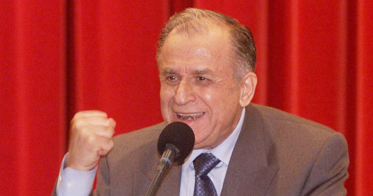 Ion Iliescu în costum vorbind la microfon, cu pumnul strâns, pe fundal roșu