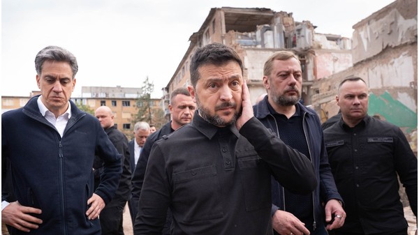 Președintele Ucrainei, Volodimir Zelenski, îmbrăcat în haine închise la culoare de stil militar, mergând alături de oficiali și gărzile sale de corp în fața unei clădiri rezidențiale grav avariate.