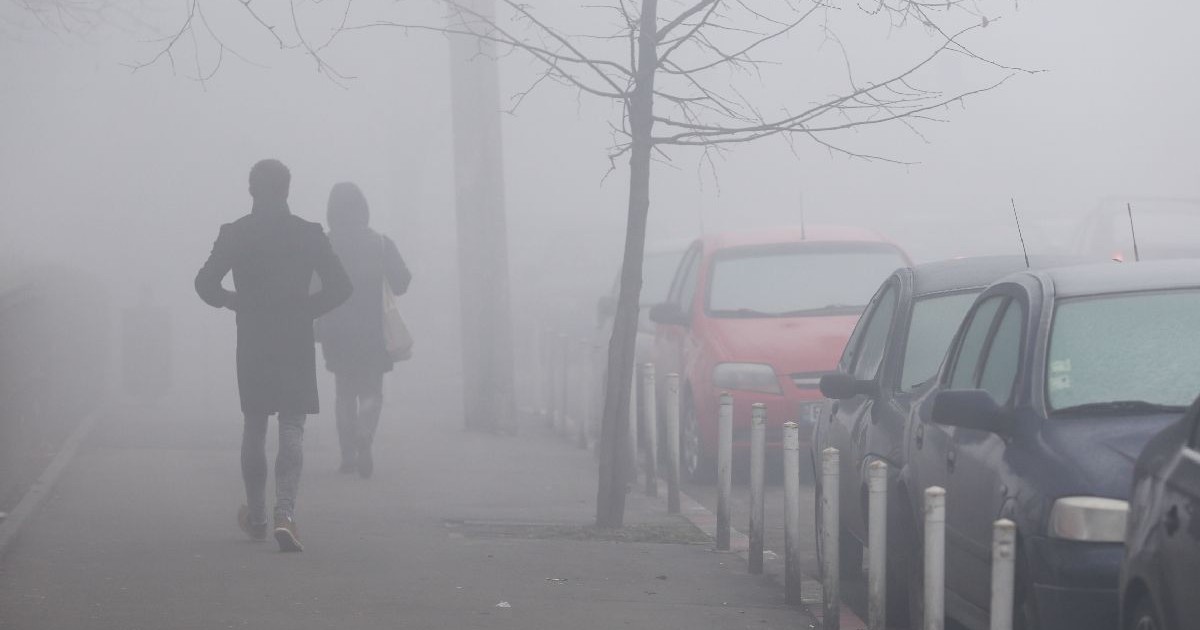 O scenă de stradă încețoșată, cu două persoane mergând pe un trotuar și mașini înghețate, parcate, pe partea dreaptă.