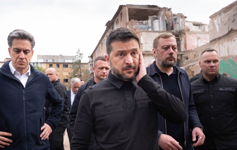 Președintele Ucrainei, Volodimir Zelenski, îmbrăcat în haine închise la culoare de stil militar, mergând alături de oficiali și gărzile sale de corp în fața unei clădiri rezidențiale grav avariate.