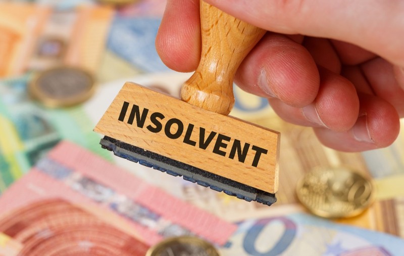 O mână care ține un ștampilă de lemn inscripționată cu cuvântul „INSOLVENT” deasupra unui fundal alcătuit din mai multe bancnote și monede euro.