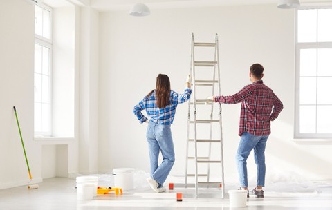 Un bărbat și o femeie priviți din spate, într-o cameră albă, aproape goală. Aceștia se află în plin proces de renovare