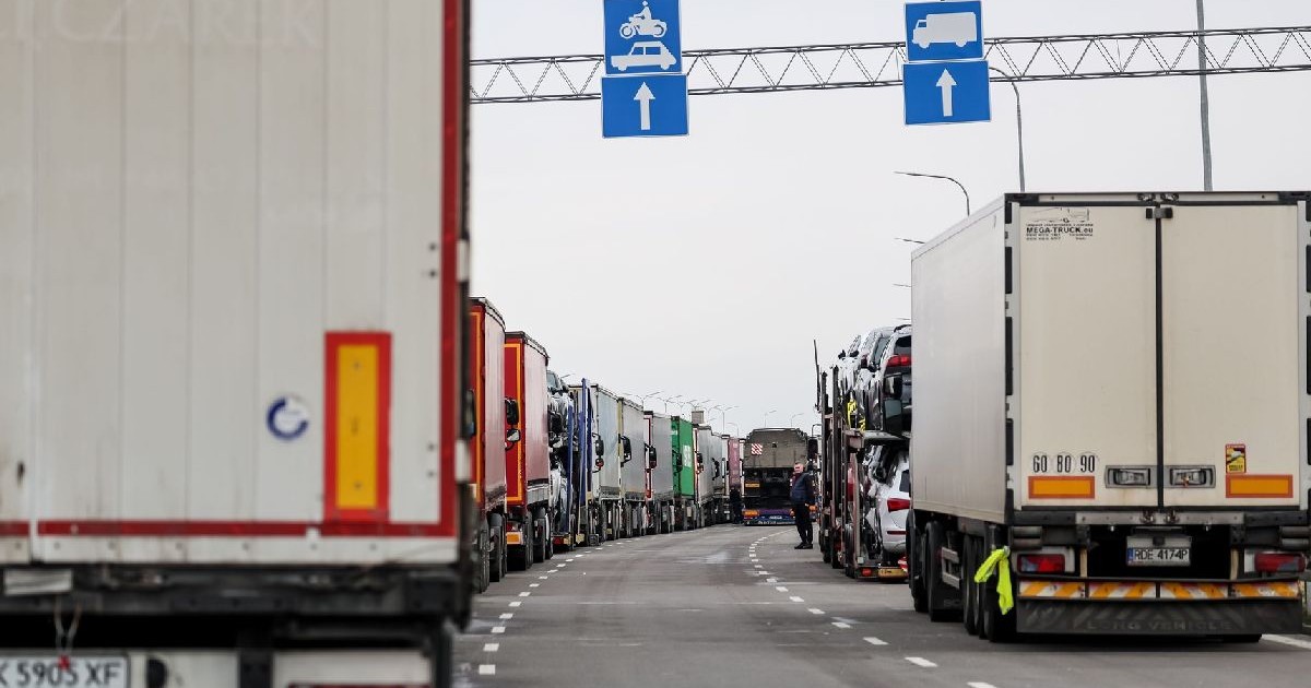 Imaginea arată o autostradă aglomerată, pe care circulă mai multe camioane mari, inclusiv un Scania alb, îndreptându-se spre un semn de control al traficului care indică virajul la stânga