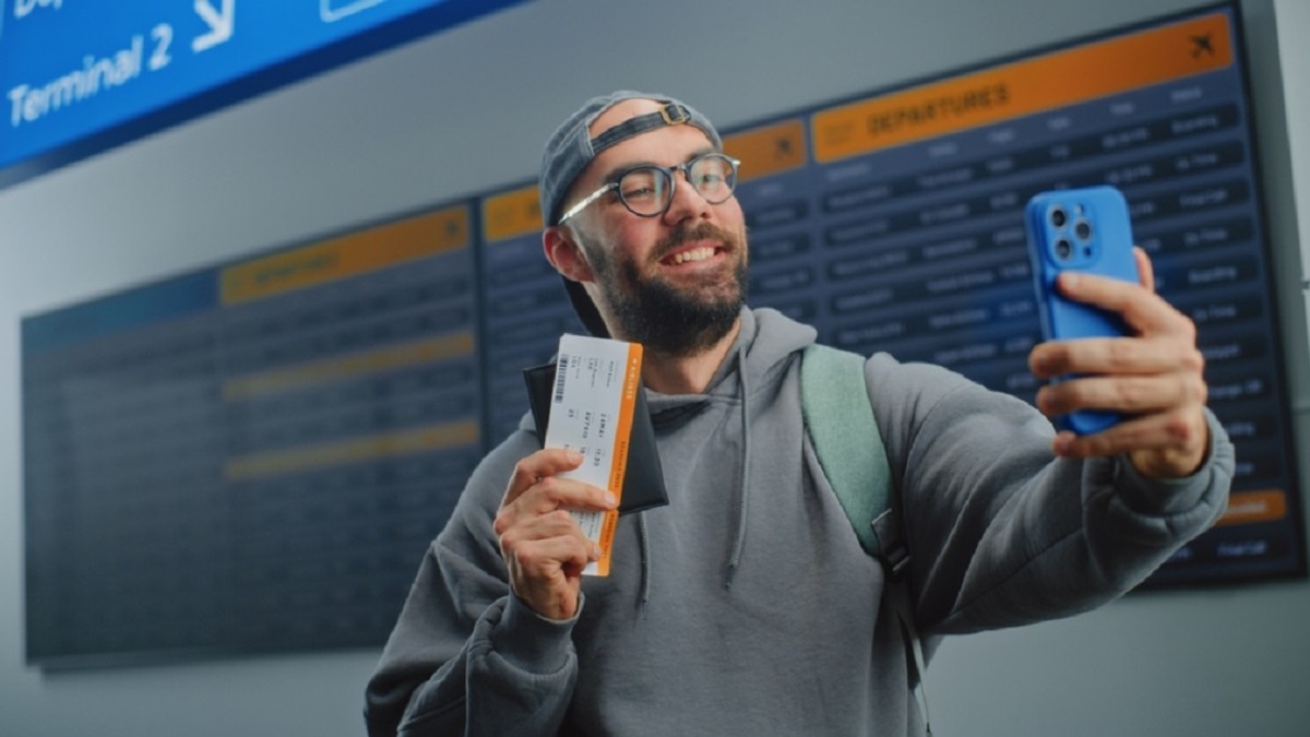 Cum să găsești bilete de avion ieftine - Un bărbat, care poartă o șapcă pe cap și ochelari de vedere, își face un selfie, într-un aeroport, ținând bucuros un bilet în mâna dreapta