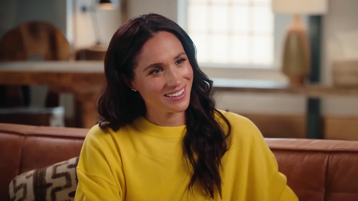 Meghan Markle, zâmbind, îmbrăcată într-un tricou galben, cu capul ușor într-o parte