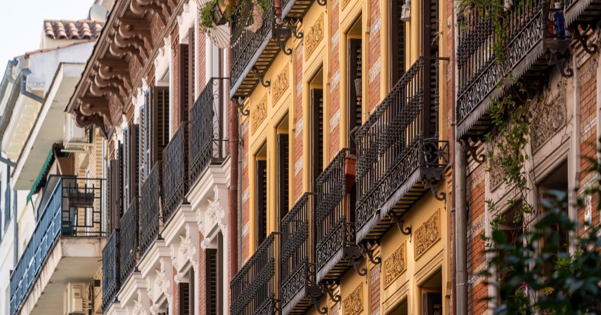 Imaginea prezintă o perspectivă în unghi strâns asupra fațadelor unor clădiri istorice din Madrid, evidențiind balcoanele decorate cu balustrade din fier forjat și ferestrele înalte cu obloane din lemn.