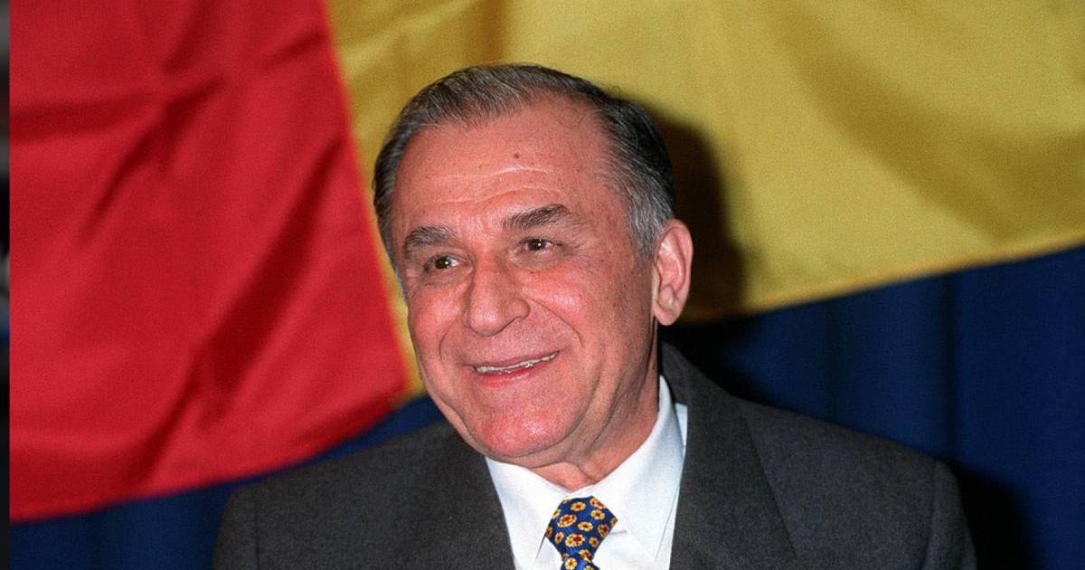 Ion Iliescu în 1994, îmbrăcat la costum cu steagul României în spatele său, este vesel și zâmbește