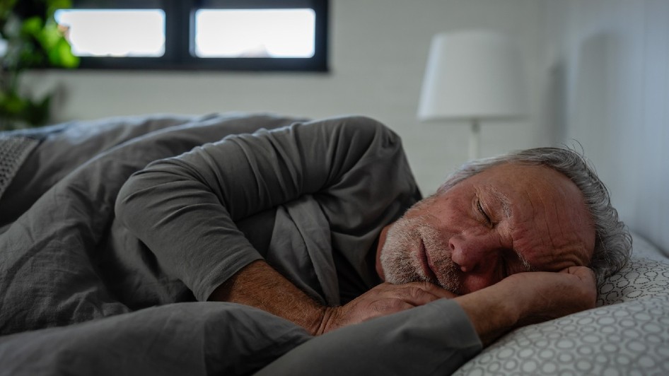Un bărbat în vârstă care doarme liniștit în pat, întins pe o parte sub o pătură gri, într-un dormitor cu lumină difuză