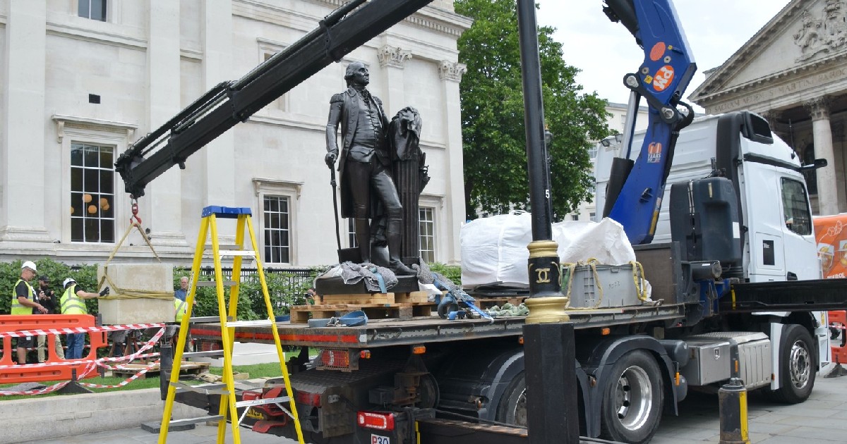 Scenă urbană în care o statuie din bronz, ce îl reprezintă pe George Washington, este transportată pe platforma unui camion. Statuia, așezată pe un suport de lemn, îl înfățișează pe Washington în picioare, îmbrăcat în haine de epocă, cu un baston în mână