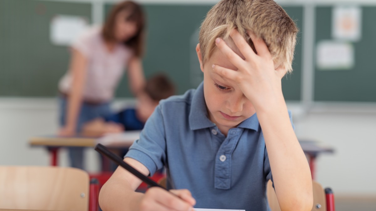 Elev de vârstă mică care stă în bancă, cu mâna în cap și se concentreaza la un test.