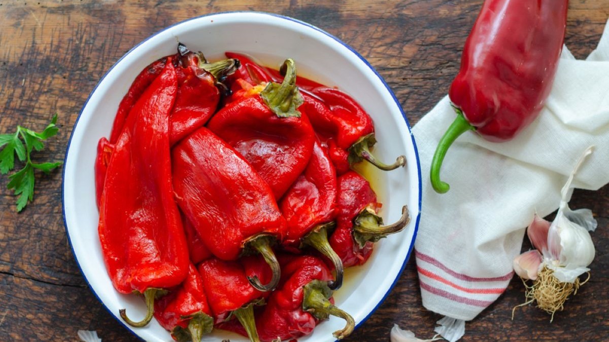 Mâini care curăță de coajă ardei capia copți dintr-un bol alb, pe un fundat de lemn