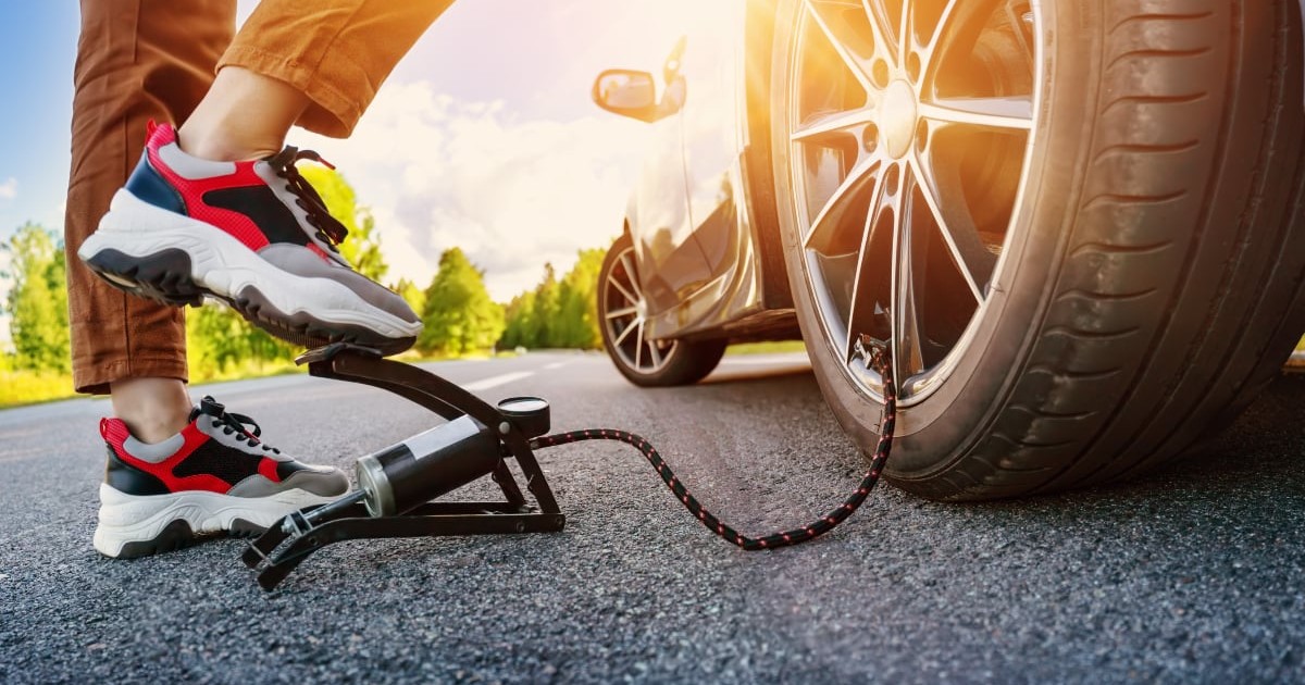 O persoană încălțată cu pantofi sport în timp ce folosește o pompă mecanică de picior pentru a umfla roata unui autoturism oprit pe marginea unui drum, sub lumina puternică a soarelui.