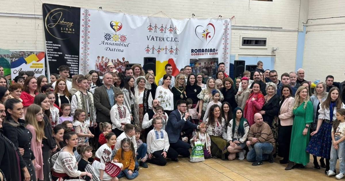 Mai multi oameni adunati intr-o incapere , stau la poza, au in spate un banner alb cu scris si motive românesti