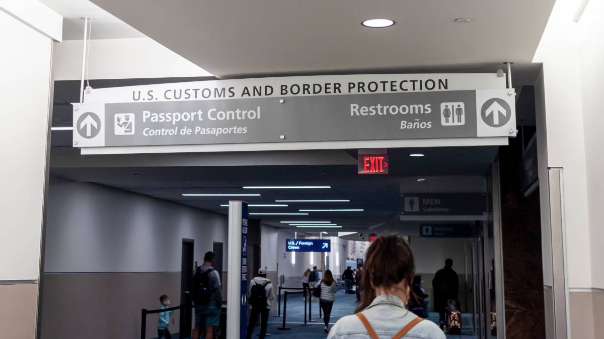 O femeie șatenă cu un ghiozdan în spate stă și așteaptă controlul de la vamă, într-un aeroport, pentru a putea intra în SUA.