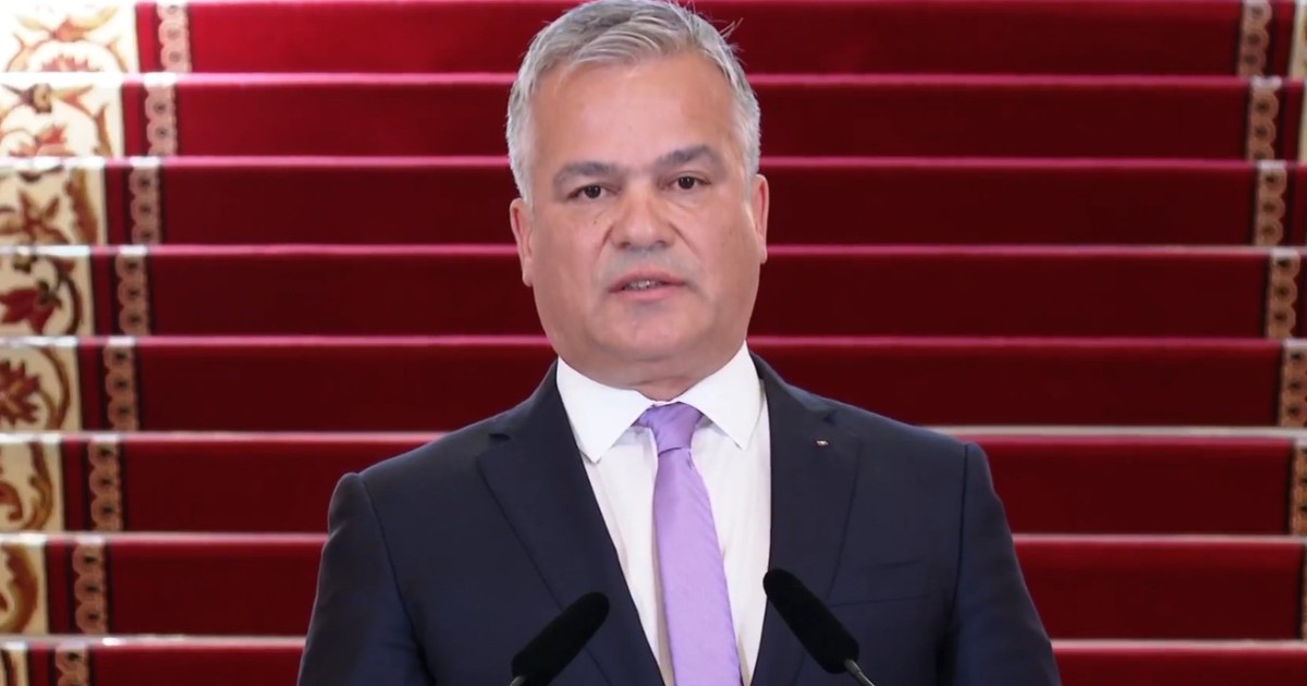 Adrian Veștea, îmbrăcat în costum închis la culoare, cămașă albă și cravată liliachie, vorbind în fața a două microfoane pe fundalul unor trepte acoperite cu o mochetă roșie