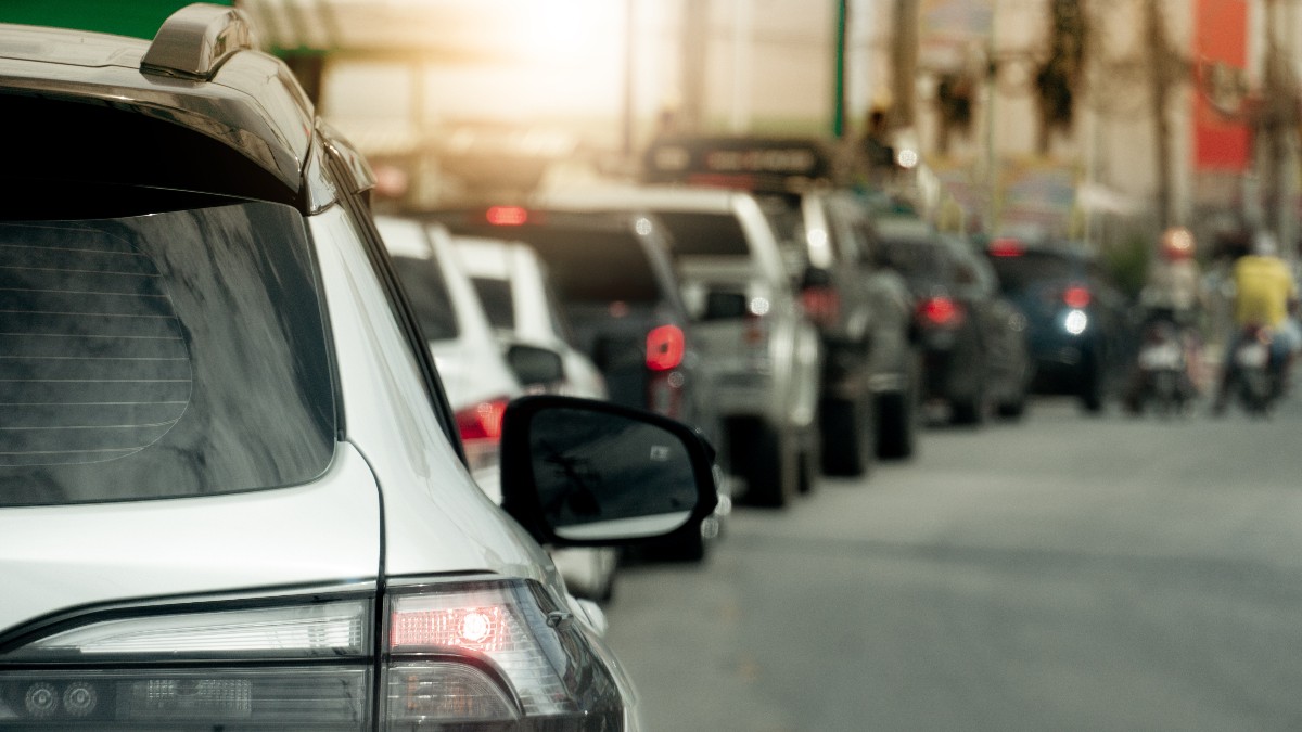 Imaginea surprinde o coloană lungă de mașini blocate în trafic, întinsă pe o stradă aglomerată. Prim-planul este ocupat de partea din spate a unui SUV alb, cu stopul aprins, semn că traficul este aproape oprit.