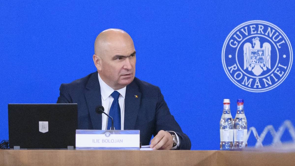 Ilie Bolojan la începutul ședinței de guvern, este îmbrăcat în costum și se uită în partea stângă.