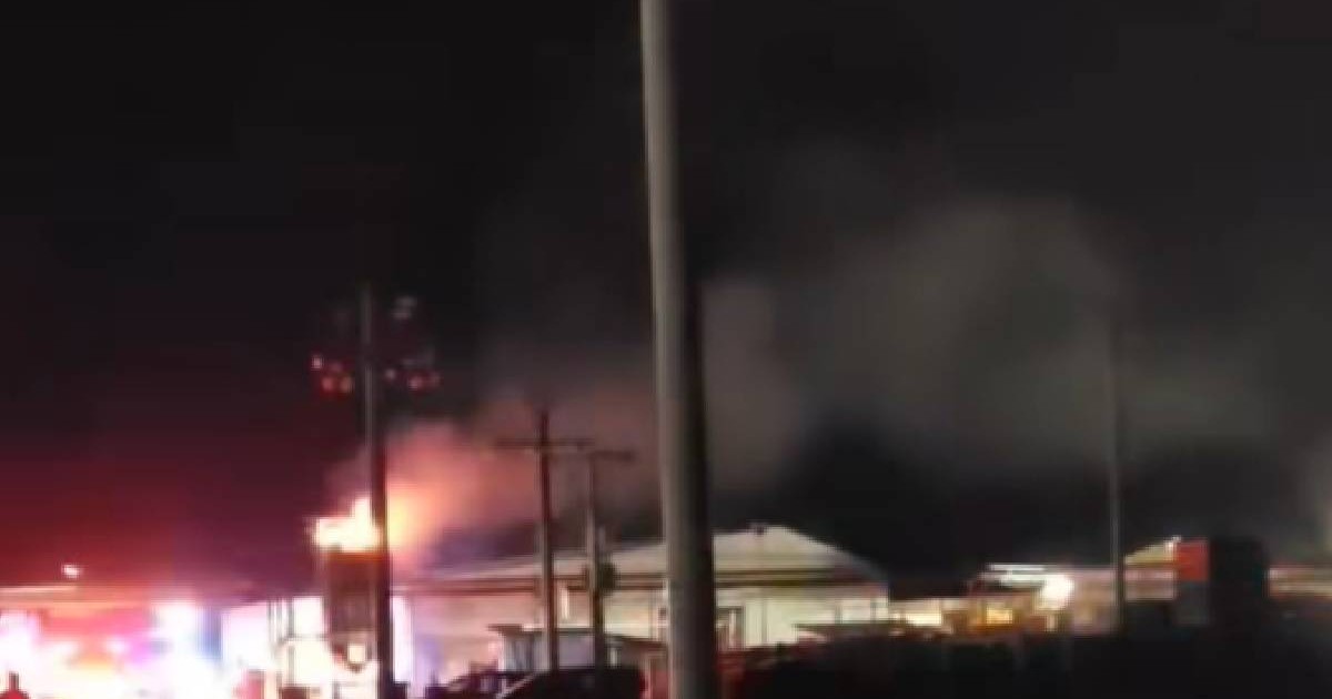 În imagine se vede un incendiu puternic pe fundal, cu oameni care privesc de pe margine încercând să rezolve situația