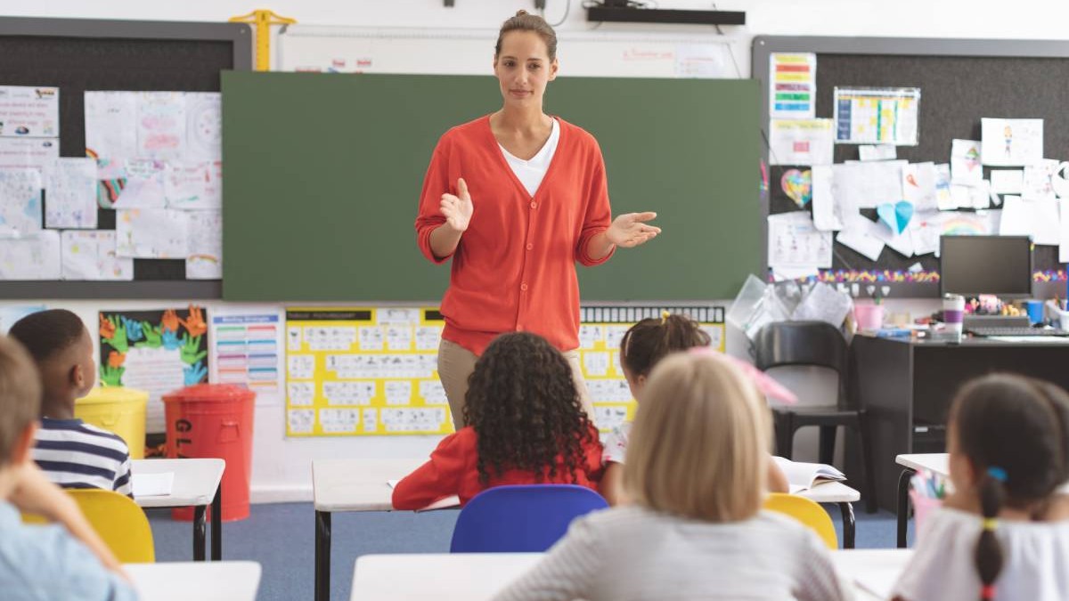 O profesoara in sala de clasa vorbește cu elevii din școala primara și le pregătește lecția, copiii sunt atenți la ceea ce le spune învățătoarea care stă în fața tablei