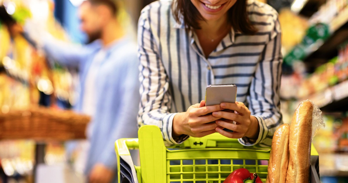 Aplicatii reduceri alimente expirare- O femeie se afla la cumparaturi si sta sprijinita pe cosul de cumparaturi in timp ce se uita in telefon