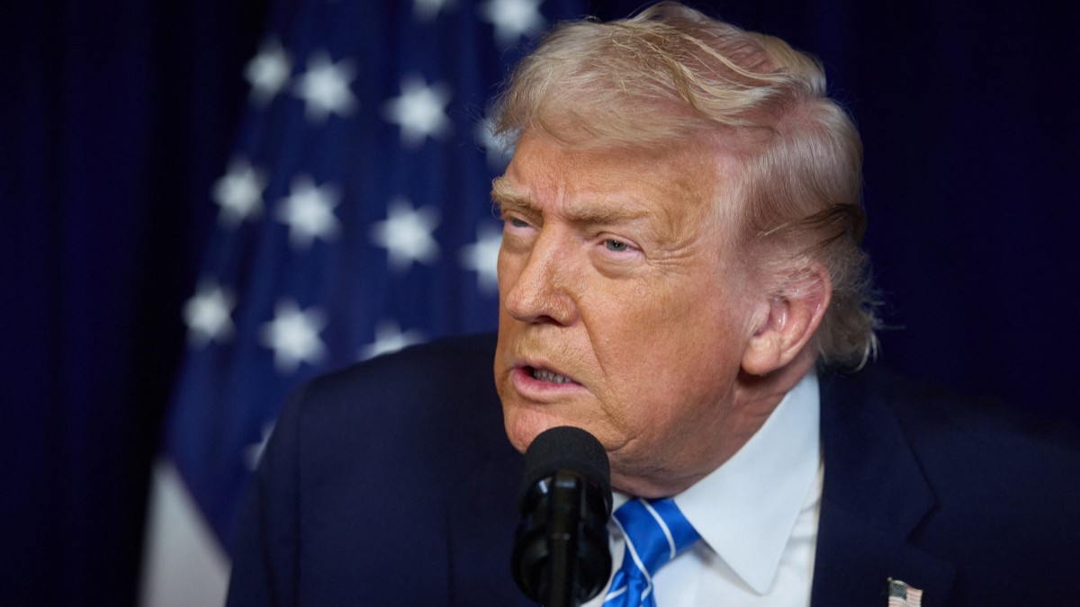 Portret Donald Trump, in sacou negru, camasa alba, cravata cu dunci albastre si insigna in piept, surprins in timp ce vorbeste la microfon, in spate se vede steagul SUA