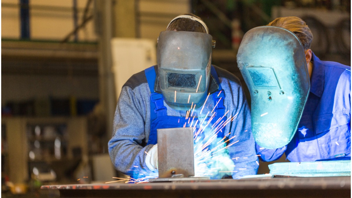 Doi muncitori echipați cu măști de protecție care rsudează într-un atelier industrial, în timp ce sar scântei luminoase.