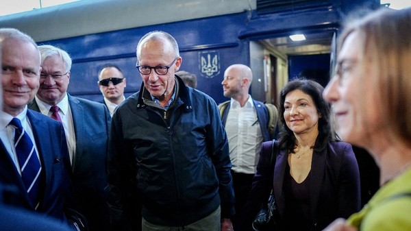 Friedrich Merz purtând o jachetă închisă la culoare și ochelari, încadrat de oficiali și însoțitori lângă un vagon de tren ucrainean.