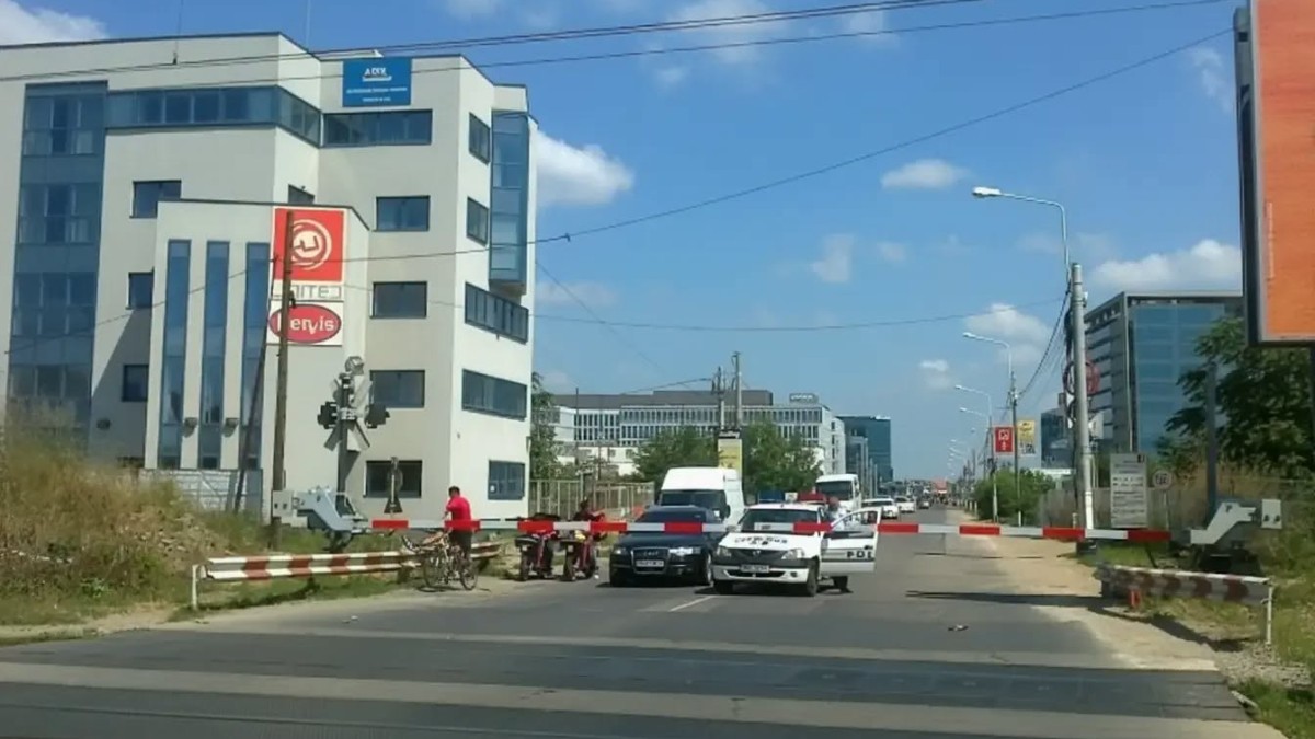 Imaginea surprinde o arteră urbană blocată de o barieră, cu mai multe mașini de poliție care controlează accesul. În fundal se observă clădiri moderne și o zonă aflată în dezvoltare, iar câțiva bicicliști așteaptă la trecerea blocată.