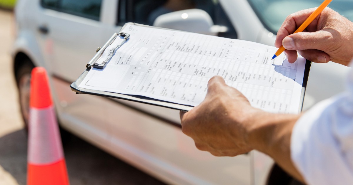 Un examinator auto care scrie pe o foaie. În fundal se observă o mașină albă.