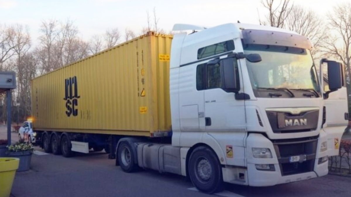 Fotografie cu un camion MAN alb cu o remorcă galbenă pe un drum asfaltat, cu copaci fără frunze în fundal.