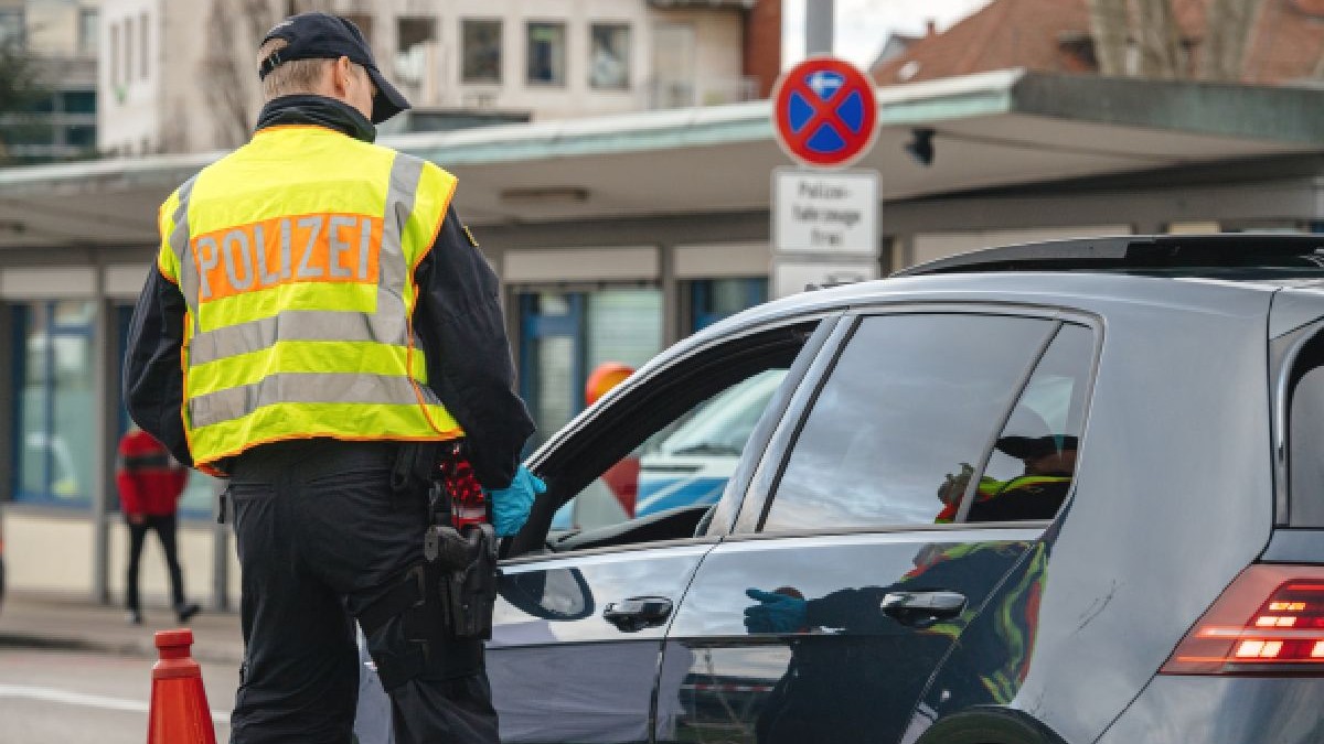 Fotografie cu un polițist german îmbrăcat într-o vestă reflectorizantă care verifică o mașină neagră, cu un semn de interdicție de parcare și pietoni în fundal.
