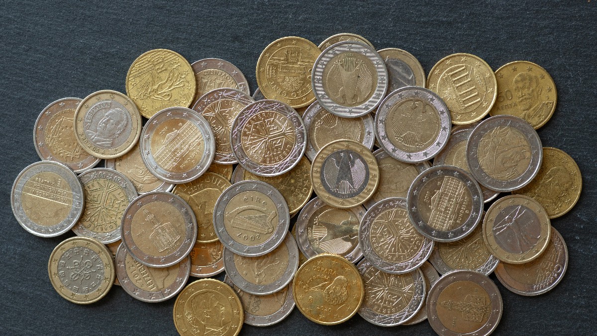 Imaginea prezintă un teanc de monede euro, de diferite valori, împrăștiate pe o suprafață închisă la culoare. Se pot observa atât monede de 1 și 2 euro, cât și piese mai mici, cu modele și simboluri specifice mai multor țări din zona euro.